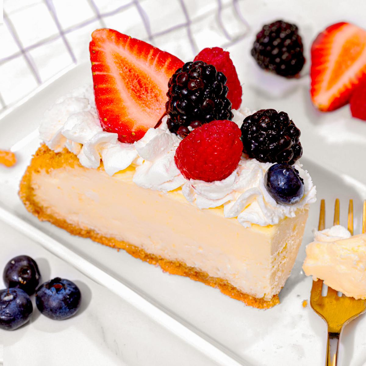 A slice of creamy cheesecake with a graham cracker crust, topped with whipped cream, fresh strawberries, blackberries, raspberries, and blueberries, served on a white rectangular plate with a gold fork.