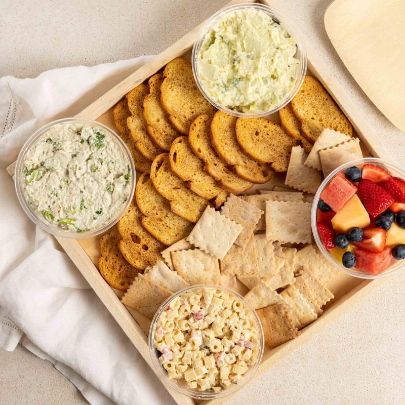 A tray of macaroni salad, fruit salad, chicken salad, crackers and carrot sticks