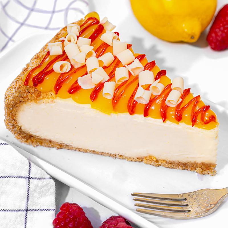 A slice of creamy lemon cheesecake with a graham cracker crust, topped with lemon curd, a raspberry drizzle, and white chocolate curls, served on a white plate with fresh raspberries and a lemon in the background.