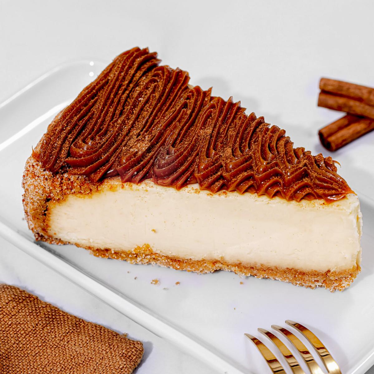 A slice of creamy cheesecake with a cinnamon sugar-coated crust, topped with piped dulce de leche and a dusting of cinnamon, served on a white plate with cinnamon sticks in the background.